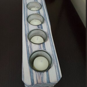 May include: A wooden candle holder with four glass candle holders. The candle holder is painted in shades of gray and white with a wood grain pattern.