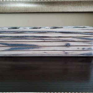 May include: A wooden box with a stacked design, featuring a light brown and black wood grain pattern. The box is made of multiple layers of wood, creating a unique and rustic look.