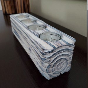May include: A white wooden candle holder with four circular openings for tealight candles. The holder has a natural wood grain pattern and a distressed finish.
