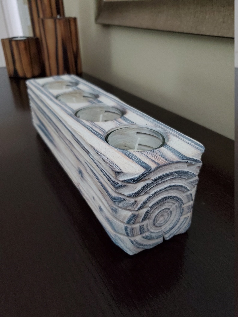 May include: A white wooden candle holder with four circular openings for tealight candles. The holder has a natural wood grain pattern and a distressed finish.