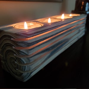 May include: A wooden candle holder with four slots for tealight candles. The holder is made of stacked wood with a natural wood grain pattern and a light brown and gray color. The candles are lit and glowing.
