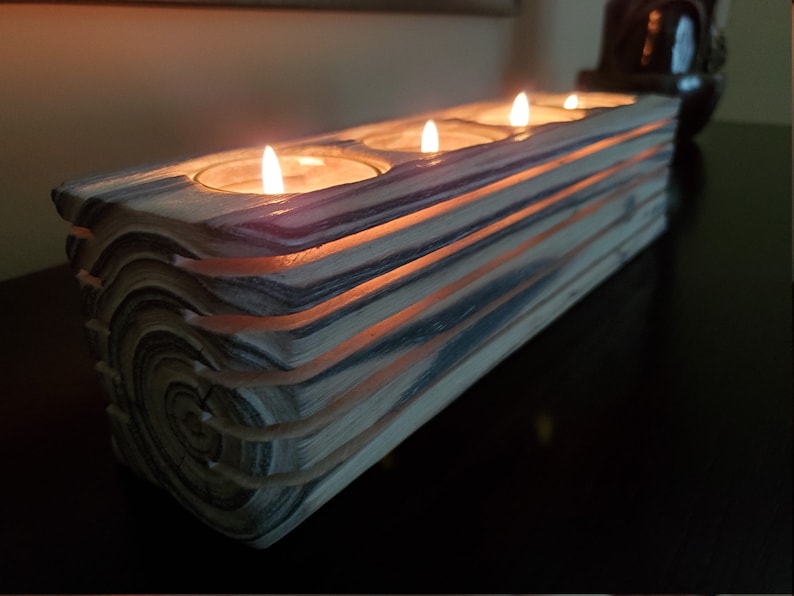 May include: A wooden candle holder with four slots for tealight candles. The holder is made of stacked wood with a natural wood grain pattern and a light brown and gray color. The candles are lit and glowing.