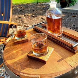 May include: A wooden barrel table with a black metal band, two glasses of amber liquid, and a clear glass decanter filled with amber liquid. The table is set with two wooden coasters.