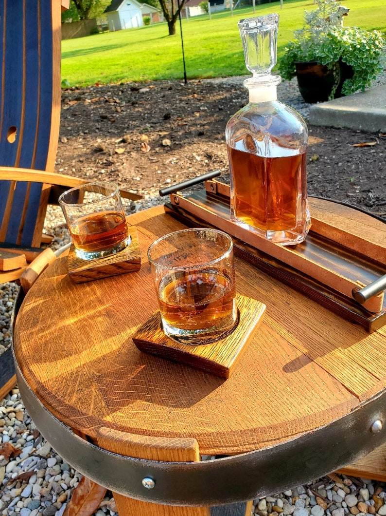 May include: A wooden barrel table with a black metal band, two glasses of amber liquid, and a clear glass decanter filled with amber liquid. The table is set with two wooden coasters.