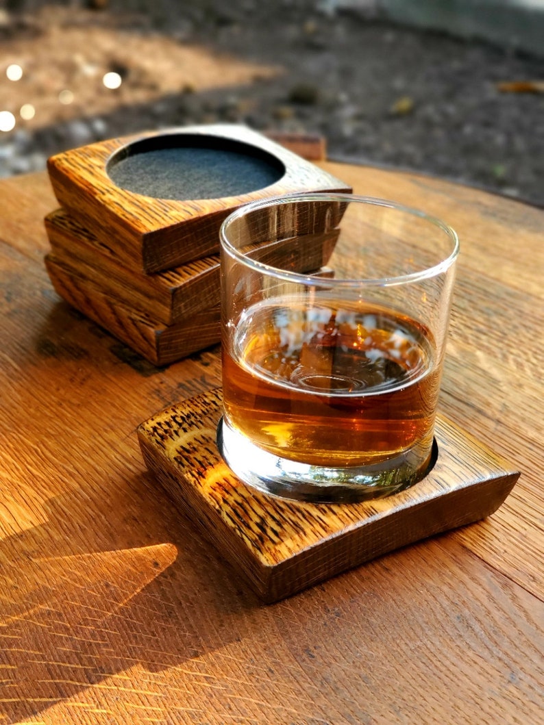 May include: A set of four wooden coasters made from reclaimed whiskey barrels, with a glass of amber liquid sitting on top of one of the coasters.