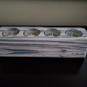 May include: A wooden candle holder with four individual slots for tealight candles. The holder is made of light-colored wood with dark grey streaks throughout.