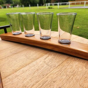 May include: A wooden serving tray with four clear glass shot glasses. The tray is made from a reclaimed barrel stave and has two black metal handles.