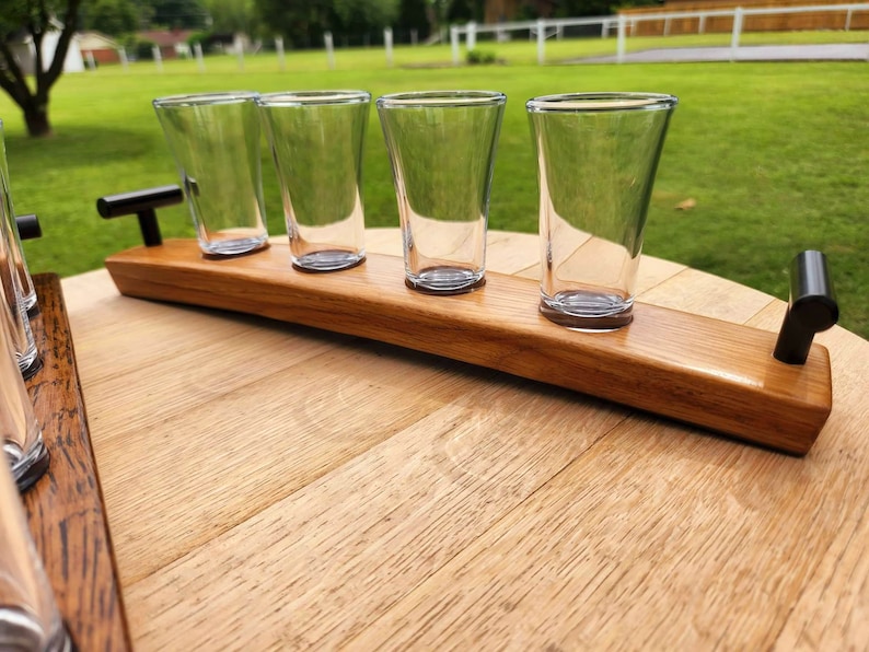 May include: A wooden serving tray with four clear glass shot glasses. The tray is made from a reclaimed barrel stave and has two black metal handles.