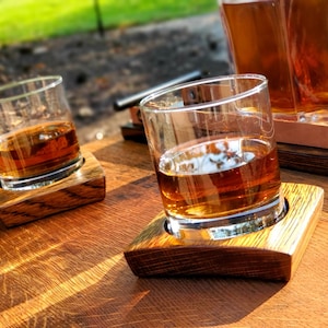 May include: Two glasses of amber-coloured liquid on wooden coasters, sitting on a wooden surface. The glasses are made of clear glass and have a rounded shape. The coasters are made of dark wood and have a rectangular shape.