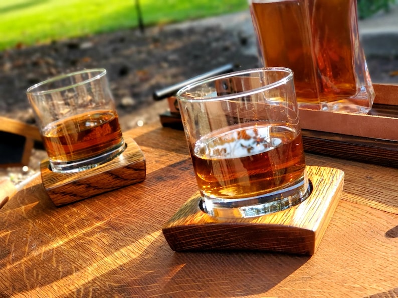 May include: Two glasses of amber-coloured liquid on wooden coasters, sitting on a wooden surface. The glasses are made of clear glass and have a rounded shape. The coasters are made of dark wood and have a rectangular shape.