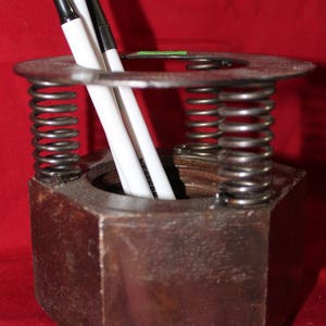 May include: A rustic metal pen holder with two springs and a hexagonal base. The holder is made from recycled materials and has a dark brown finish. Two pens are in the holder, one white and one black.