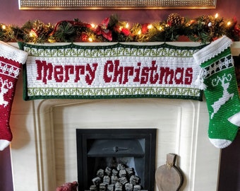 May include: A white fireplace mantel decorated with a green garland and two crocheted Christmas stockings. The stockings are red and green with a reindeer design and the text "Merry Christmas" is embroidered on the banner.