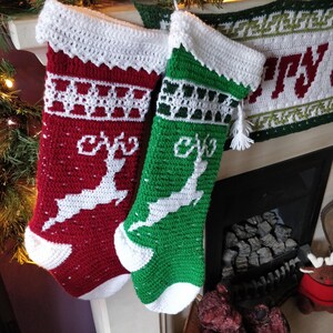 May include: Two crocheted Christmas stockings with white trim and reindeer designs. One stocking is red with white snowflakes and the other is green with white snowflakes.