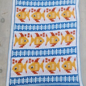 K&ouml;nnte beinhalten: Eine wei&szlig;e H&auml;keldecke mit einem blau-wei&szlig;en gestreiften Rand. Die Decke zeigt ein sich wiederholendes Muster aus gelben Fischen mit orangefarbenen Flossen und schwarzen Augen.