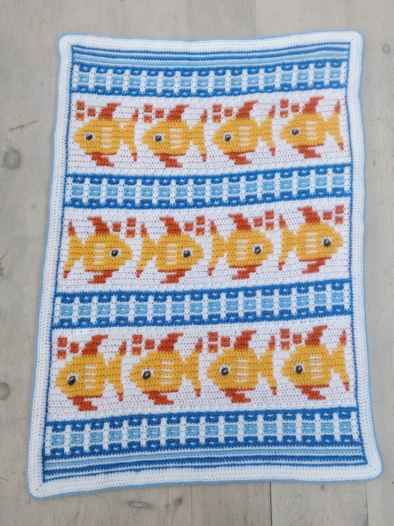 K&ouml;nnte beinhalten: Eine wei&szlig;e H&auml;keldecke mit einem blau-wei&szlig;en gestreiften Rand. Die Decke zeigt ein sich wiederholendes Muster aus gelben Fischen mit orangefarbenen Flossen und schwarzen Augen.