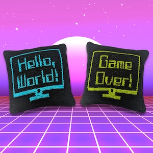 Op de afbeelding: Twee zwarte gehaakte kussens met een gepixelde computerscherm-ontwerp. Het kussen aan de linkerkant heeft de tekst "Hello, World!" in blauw. Het kussen aan de rechterkant heeft de tekst "Game Over!" in groen.