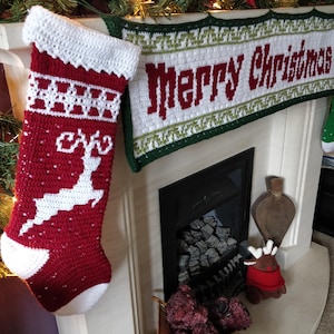 May include: A red and white crocheted Christmas stocking with a white reindeer design hangs on a fireplace mantel. Above the stocking is a white and green crocheted banner that reads "Merry Christmas".