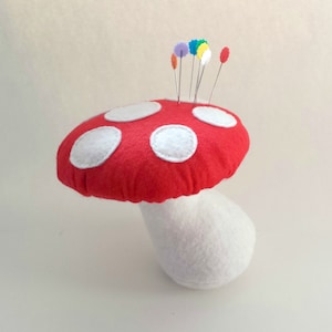 Puede incluir: Un hongo de fieltro rojo con lunares blancos y un tallo de fieltro blanco. El hongo tiene alfileres de colores clavados en la parte superior.