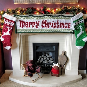 May include: A white fireplace mantel with a crocheted banner that reads "Merry Christmas". The banner is flanked by two crocheted stockings, one red and one green, with reindeer designs. A fireplace with a black grate and a pile of logs is visible behind the banner. Two stuffed animals, a brown bear and a reindeer, are sitting on the floor in front of the fireplace.