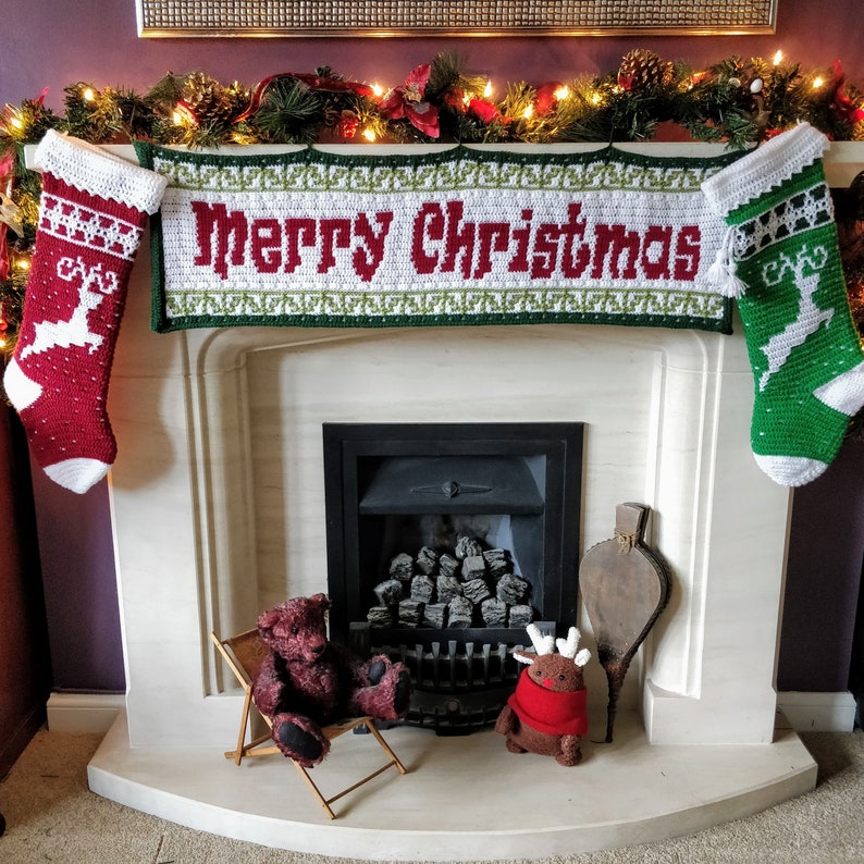 May include: A white fireplace mantel with a crocheted banner that reads "Merry Christmas". The banner is flanked by two crocheted stockings, one red and one green, with reindeer designs. A fireplace with a black grate and a pile of logs is visible behind the banner. Two stuffed animals, a brown bear and a reindeer, are sitting on the floor in front of the fireplace.