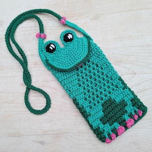 May include: A handmade crochet frog-shaped phone case in shades of teal and green. The case features a frog face with black and white eyes, a pink tongue, and a long green strap. Pink accents are on the strap and feet.