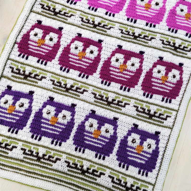 May include: Crocheted blanket featuring a repeating pattern of owls in shades of purple and pink. The owls have white eyes and orange beaks, set against a white background with green and brown decorative elements. The blanket is made of soft yarn.