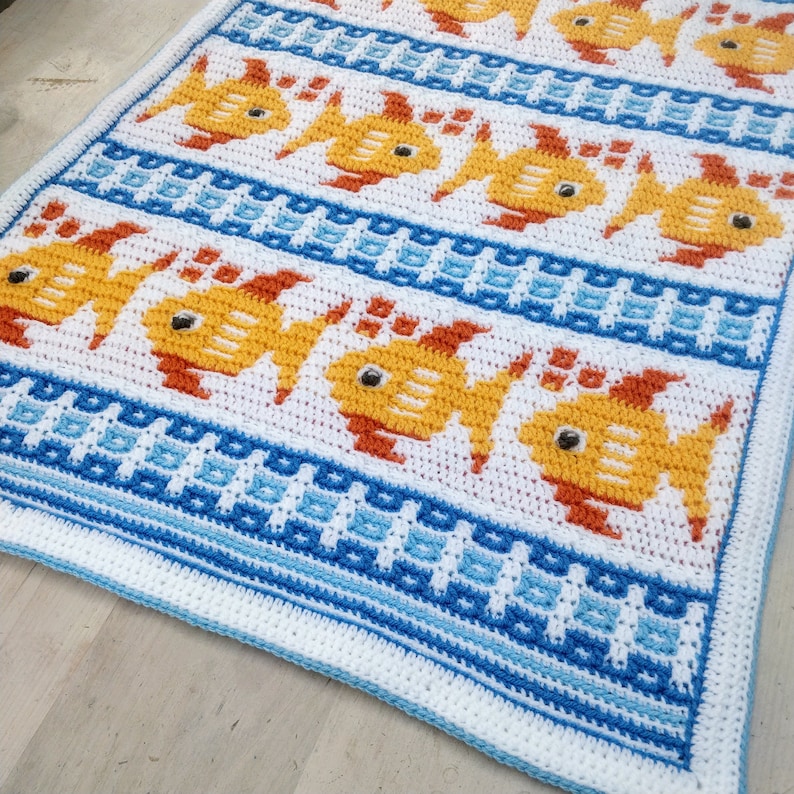 K&ouml;nnte beinhalten: Eine geh&auml;kelte Babydecke mit wei&szlig;em Hintergrund und einem Muster aus orangefarbenen und gelben Fischen, die in blauen und wei&szlig;en Wellen schwimmen.