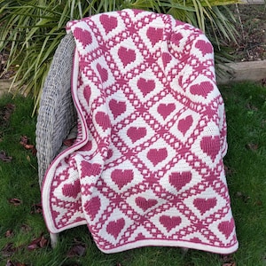 May include: A handmade, cream-colored blanket with a repeating pattern of pink hearts. The blanket is draped over a wicker chair on a grassy surface. The hearts are arranged in a diamond pattern, creating a cozy and decorative textile.