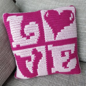Könnte beinhalten: Ein rosa und weißes Häkelkissen mit dem Wort "LOVE" in Pixelform.