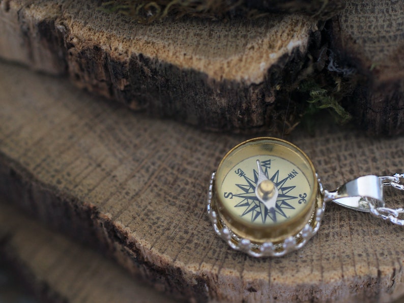 Peut inclure: Un collier avec un pendentif en forme de boussole dor&eacute;e et une cha&icirc;ne en argent. Le cadran de la boussole a des marques noires pour les points cardinaux et une aiguille en argent.