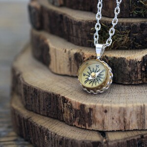 Peut inclure: Un collier en cha&icirc;ne argent&eacute;e avec un pendentif en forme de boussole. La boussole est ronde et a un cadran de couleur or avec des marques noires. La boussole est pos&eacute;e sur une pile de rondins de bois.