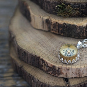 Peut inclure: Un collier en cha&icirc;ne argent avec un pendentif en forme de boussole. La boussole est ronde et a une bordure dor&eacute;e. La boussole est pos&eacute;e sur une pile de tranches de bois.