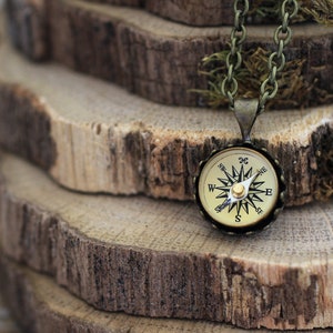 Peut inclure: Un collier avec un pendentif en forme de boussole dor&eacute;e et une cha&icirc;ne noire. La boussole a un cadran blanc avec des marques noires et une aiguille dor&eacute;e. Le collier est pos&eacute; sur une pile de rondins de bois.
