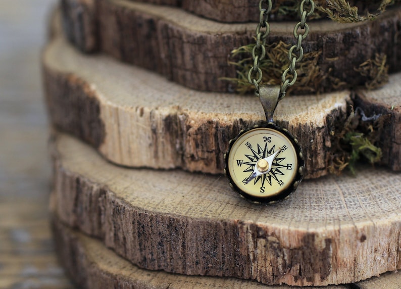 Peut inclure: Un collier avec un pendentif en forme de boussole dor&eacute;e et une cha&icirc;ne noire. La boussole a un cadran blanc avec des marques noires et une aiguille dor&eacute;e. Le collier est pos&eacute; sur une pile de rondins de bois.