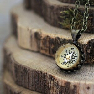 Peut inclure: Un collier avec un pendentif en forme de boussole dor&eacute;e. Le cadran de la boussole a des marques noires et une aiguille dor&eacute;e. Le collier est sur un fond en bois.