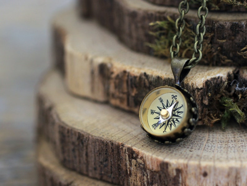 Peut inclure: Un collier avec un pendentif en forme de boussole dor&eacute;e. Le cadran de la boussole a des marques noires et une aiguille dor&eacute;e. Le collier est sur un fond en bois.