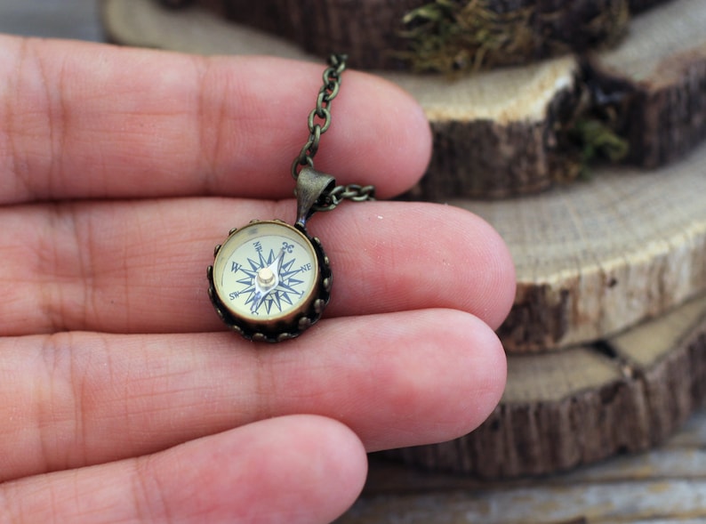 Peut inclure: Un collier avec un pendentif en forme de boussole ronde en bronze. Le cadran de la boussole est blanc avec des marques noires et une aiguille dor&eacute;e.