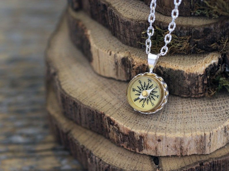 Peut inclure: Un collier en cha&icirc;ne argent avec un pendentif en forme de boussole. La boussole est ronde et a un cadran de couleur or avec des marques noires. La boussole est plac&eacute;e sur un fond de rondins de bois empil&eacute;s.