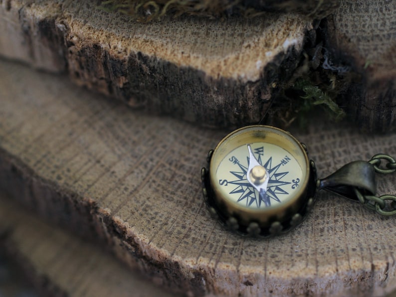 Peut inclure: Un petit pendentif en forme de boussole d'aspect antique avec une finition dor&eacute;e. Le cadran de la boussole pr&eacute;sente un motif noir et blanc avec une aiguille argent&eacute;e pointant vers le nord. Le pendentif est pos&eacute; sur une surface en bois rustique.