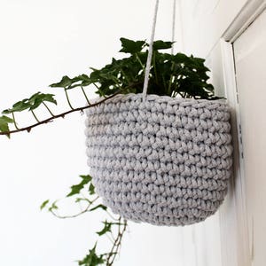 Puede incluir: Un macetero colgante de crochet gris con una planta verde en su interior. El macetero está colgado de una pared blanca.