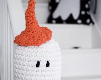 May include: A white crocheted ghost figure with a textured surface, featuring two small black oval eyes and a pointed orange crocheted hat resting on top. The ghost is crafted from thick yarn and has a cylindrical shape with a rounded top.