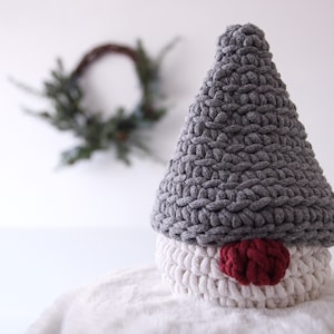 May include: A gray crocheted gnome with a white crocheted base and a red felt nose. The gnome is sitting on a white surface.