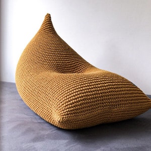 Può includere: Una grande poltrona a sacco di colore giallo senape in maglia. La poltrona è realizzata con filato spesso e ha una forma rilassata e confortevole.