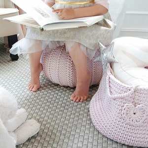 Puede incluir: Una ni&ntilde;a peque&ntilde;a est&aacute; sentada en un puf de punto rosa mientras lee un libro. Una cesta de punto rosa con un dise&ntilde;o de coraz&oacute;n blanco est&aacute; en el suelo junto al puf. Un conejito de peluche blanco est&aacute; en el suelo en primer plano.