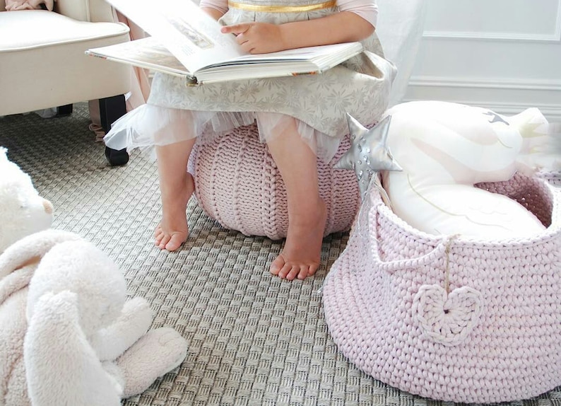 Puede incluir: Una ni&ntilde;a peque&ntilde;a est&aacute; sentada en un puf de punto rosa mientras lee un libro. Una cesta de punto rosa con un dise&ntilde;o de coraz&oacute;n blanco est&aacute; en el suelo junto al puf. Un conejito de peluche blanco est&aacute; en el suelo en primer plano.