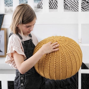 May include: A young person is holding a large, round, mustard yellow knitted pouf. The pouf has a textured, ribbed surface.