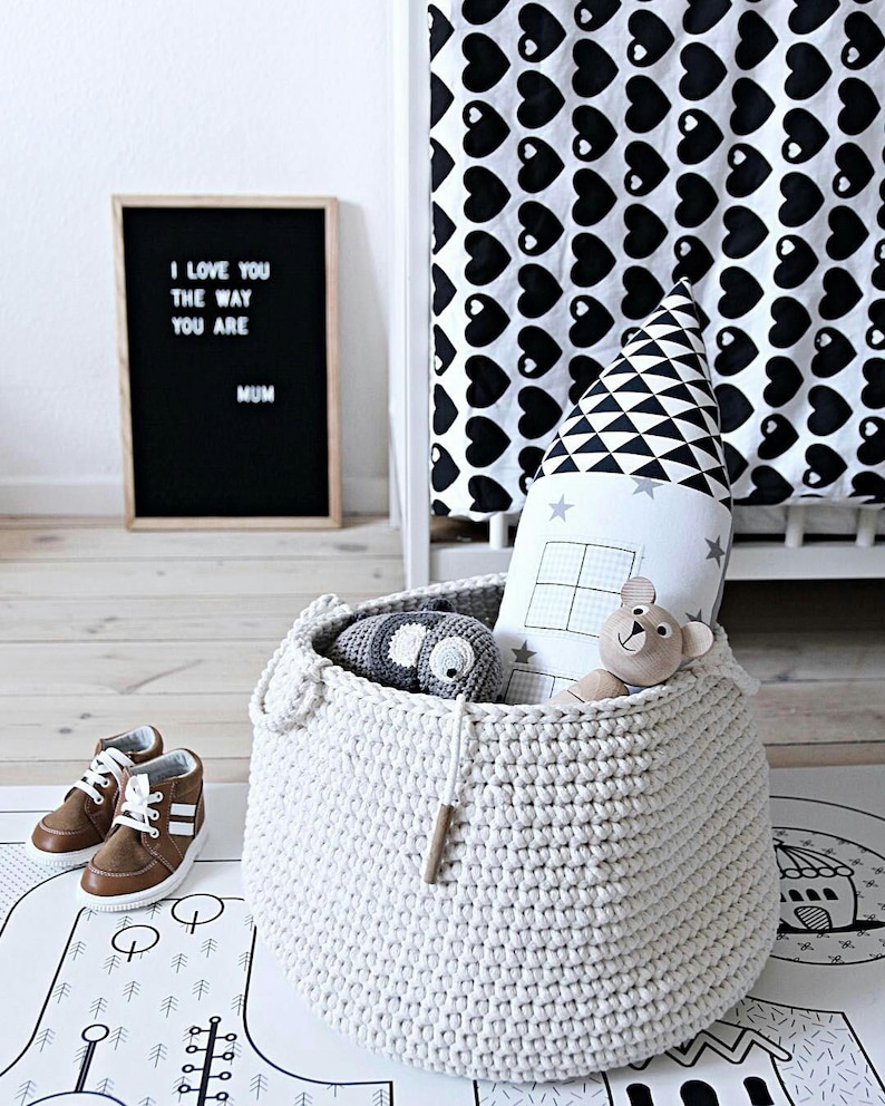 Puede incluir: Una cesta de almacenamiento de crochet blanca llena de juguetes de peluche, incluyendo un elefante gris, una estrella blanca y un oso marr&oacute;n. La cesta est&aacute; sentada en una alfombra de juego negra y blanca con un dise&ntilde;o geom&eacute;trico.