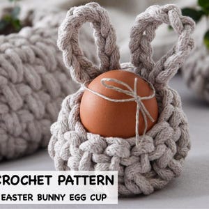May include: A light grey crocheted Easter bunny egg cup holding a brown egg tied with twine. The bunny ears form a handle. The text "CROCHET PATTERN EASTER BUNNY EGG CUP" is at the bottom.
