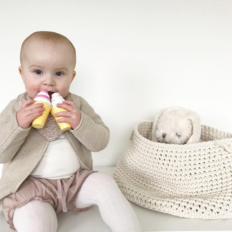 Puede incluir: Un beb&eacute; con un c&aacute;rdigan beige y un babero estampado en rosa y blanco sostiene dos conos de helado de juguete. Una cesta de crochet blanca con un conejito de peluche blanco en su interior se encuentra sobre una superficie blanca.