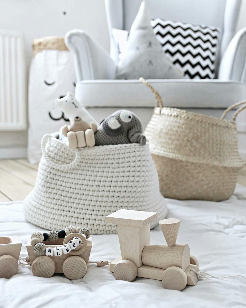 Puede incluir: Una cesta de almacenamiento de crochet blanca llena de animales de peluche, un tren de juguete de madera con una etiqueta de nombre que dice "ALBERT" y un collar de cuentas. La cesta est&aacute; sentada sobre una alfombra blanca frente a un sill&oacute;n blanco con una almohada de chevron blanco y negro.